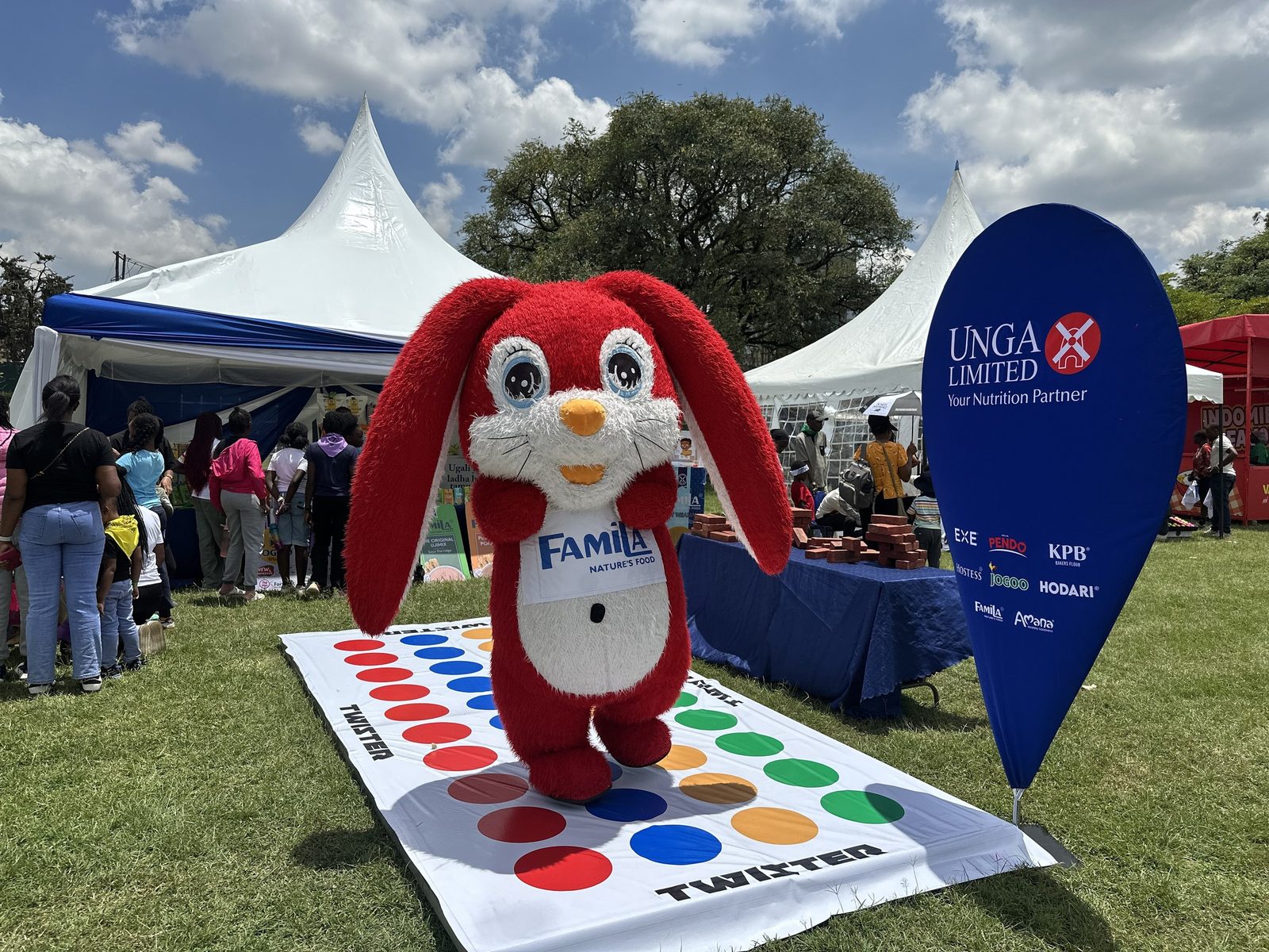 A brand mascot standing on an oversized Twister mat laid on grass beside a branded stand